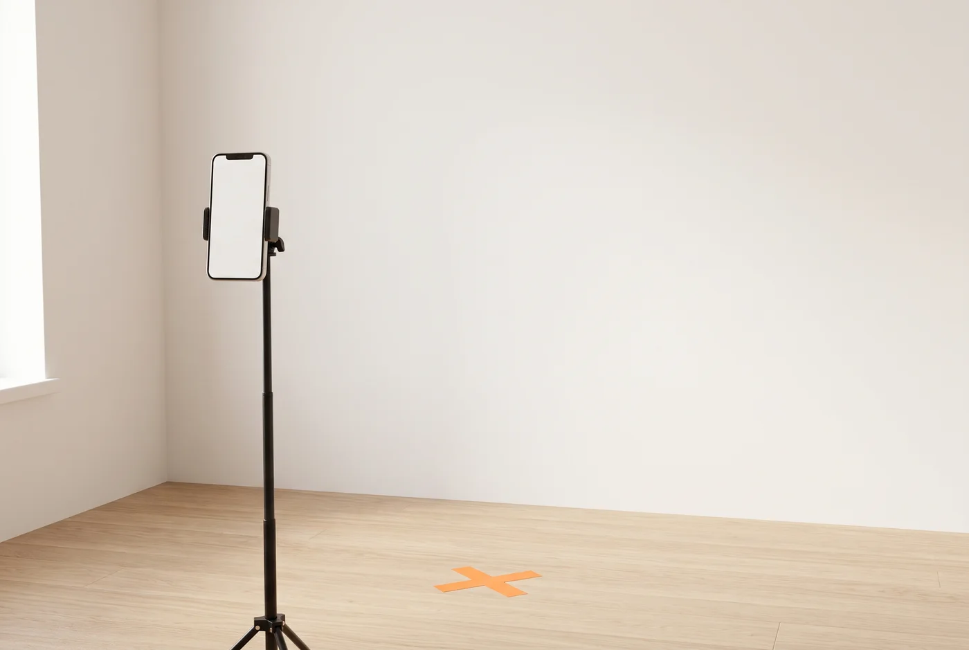 A phone on a tripod in a bright, empty room corner, aimed at a plain off-white wall with an orange tape X on the wood floor marking where to stand