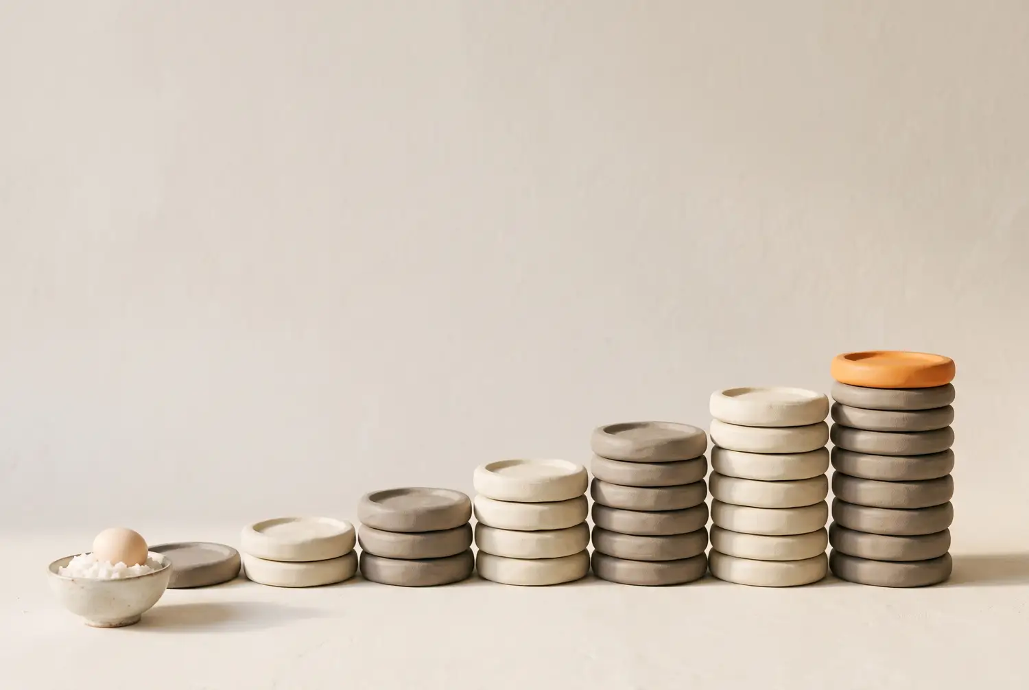 Soft clay illustration of weight plates stacked into a gentle staircase, each stack only slightly taller than the last, ending in an orange plate, with a small bowl of rice and an egg at the start